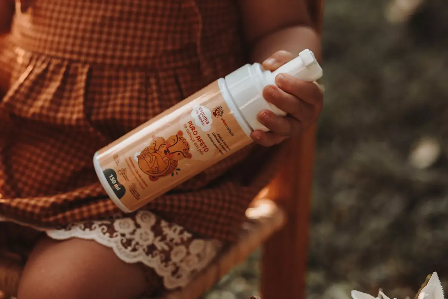 Close na mão de uma criança segurando a espuma de banho da Afeto Natural Menina sentada em banco no jardim, segurando os produtos da Afeto Natural - Just Real Moms