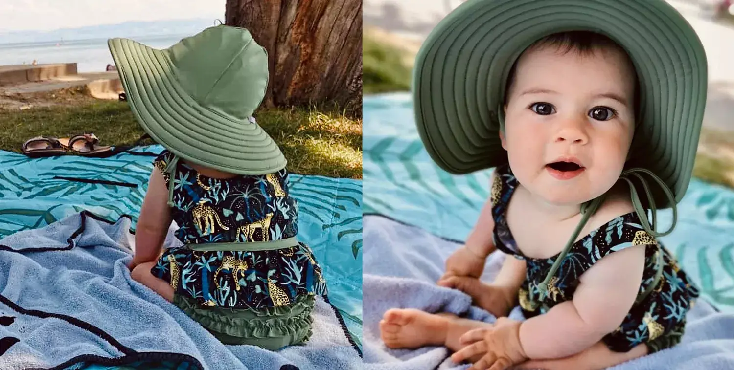 Bebe sentada a beira da praia em gramado com roupa da Mimor Kids e chapéu grande verde